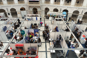 Große Besuchermenge auf einer internationalen Fachmesse mit Ständen, Bannern, Tischen, Stühlen und Ballons in einem Raum mit Bogengängen, Säulen und einem Bildschirm im Hintergrund.