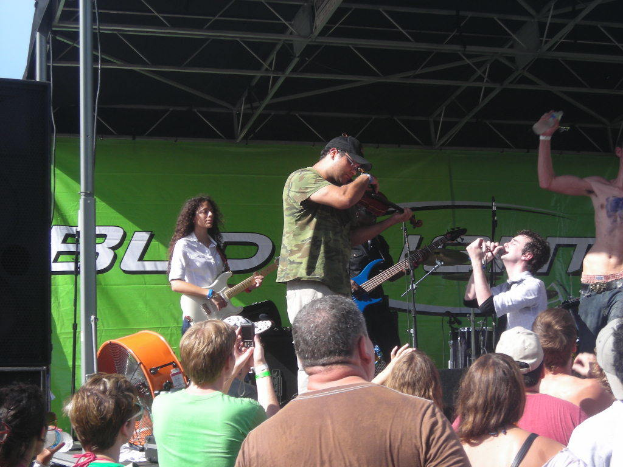 Eine Gruppe von Musikern spielt verschiedene Instrumente auf einer Bühne mit einem großen schwarzen Lautsprecher links daneben, während das Publikum davor steht und die Vorstellung genießt, mit einem grünen Banner im Hintergrund.