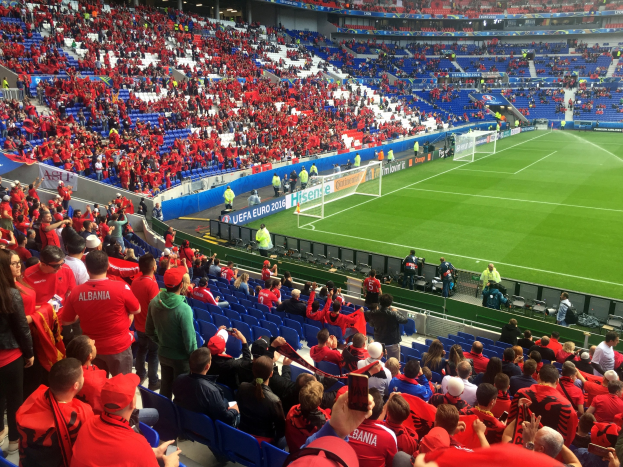 Eine große Menschenmenge in einem Stadion schaut einem Fussballspiel zu, mit Menschen auf Sitzen und Stehplätzen, Fahnen, einem Torpfosten und Deckenbeleuchtung.