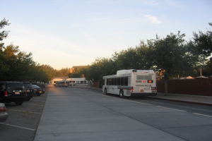 Ein Shuttle-Bus fährt auf einer Straße mit parkenden Autos, Bäumen und Gebäuden auf beiden Seiten unter einem klaren blauen Himmel.