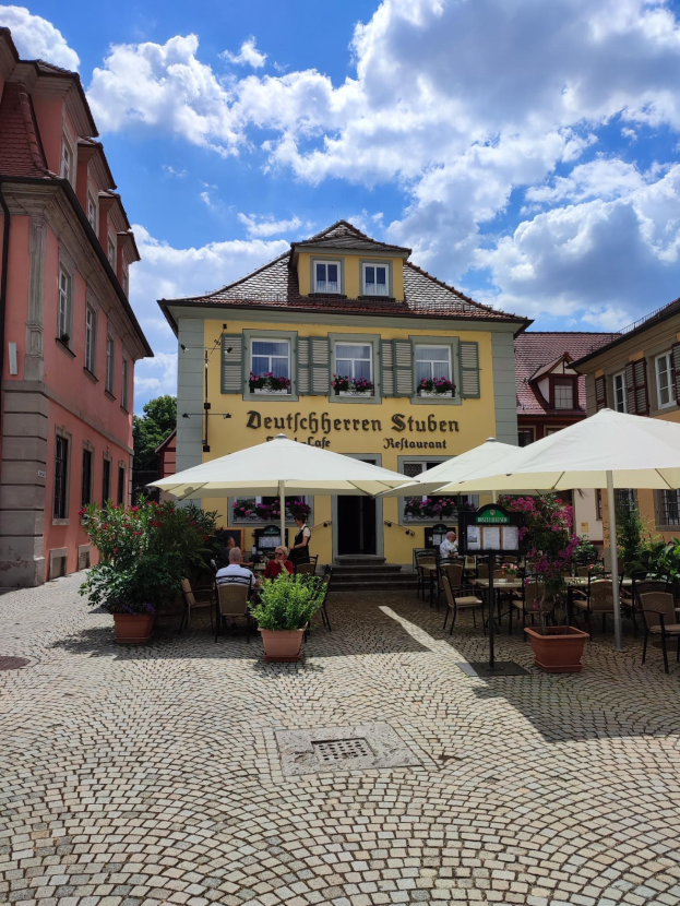 Außensitzbereich mit Tischen, Sonnenschirmen und einigen Menschen vor dem gelben Gebäude der Deutschen Stuben Restaurant unter einem blauen und weißen Himmel.