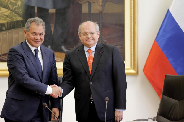 Zwei Männer im Blazer geben sich die Hand vor Mikrofonen, mit einem Stuhl, einer Flagge und einem gerahmten Wandbild im Hintergrund.