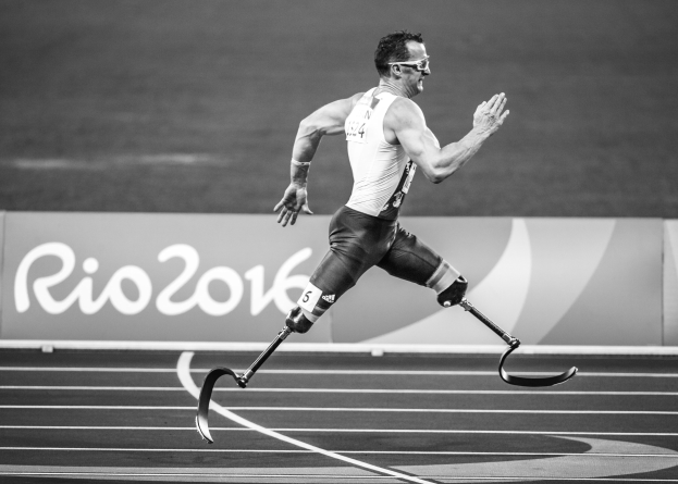 Ein Mann mit einer Prothese läuft auf einer Laufbahn mit einer Tafel "Rio 2016 Paralympische Spiele" im Hintergrund, in Schwarz-Weiß.
