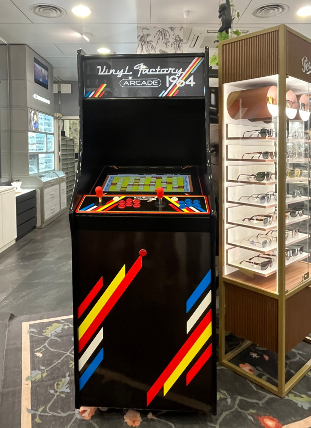 Ein Geschäftsinnenraum mit einem vintage Arcade-Automaten in der Mitte, einem Glasvitrinenschrank auf der rechten Seite und retro-stiliger Beleuchtung mit Schränken und einem Fernseher im Hintergrund.