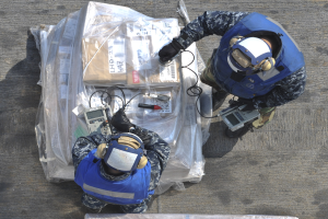 Zwei Männer in blauen Uniformen und Helmen arbeiten an einem großen Drohnen, halten Werkzeuge, mit einer Plastikabdeckung auf dem Boden.
