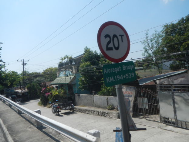 Tempolimitschild am Straßenrand mit Fahrzeugen, Geländer, Bäumen, Gebäuden, Strommasten mit Drähten und einem bewölkten Himmel im Hintergrund.