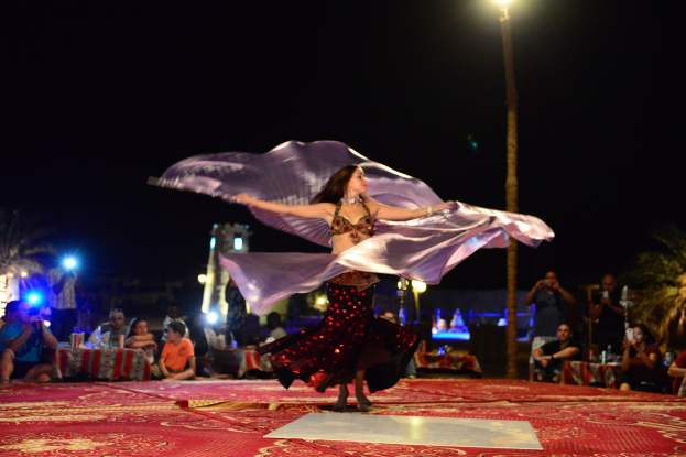 Eine Frau in einem Bauchtanzkostüm tanzt auf einem roten Teppich, umgeben von einer Zuschauermenge mit Kameras vor einem Hintergrund aus Bäumen, Lichtern, einem Pfahl und Himmel.