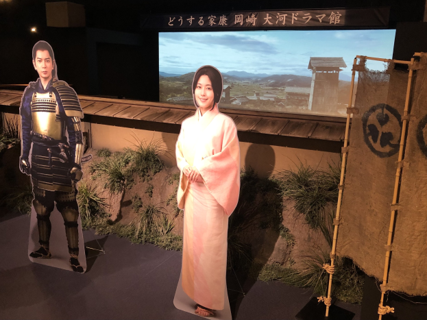 Eine Frau in einem rosa Kimono und ein Mann in einem Samurai-Kostüm, die zusammen lächeln, in einer Szene aus einem japanischen Film mit Pflanzen, einem Bildschirm und Lampen im Hintergrund.