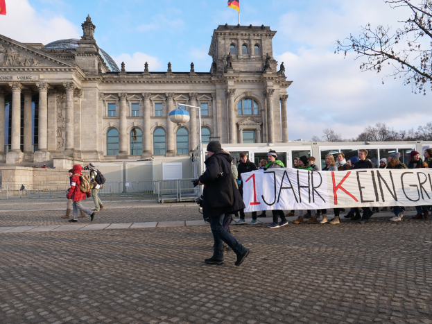 Eine Gruppe von Menschen marschiert vor dem Reichstaggebäude in Berlin, eine Fahne haltend, mit Säulen, Fenstern, Bögen, Statuen und Flaggen geschmückt, umgeben von einem Metallzaun, einer Straßenlaterne, Bäumen und einem bewölkten Himmel im Hintergrund.