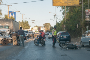 Gruppe von Menschen um ein verunglücktes Motorrad auf der Straße mit mehreren Fahrzeugen, darunter ein Lastwagen, und Hintergrundelementen wie Bäumen, Pfählen, Lampen, Schildern und Himmel.