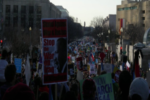 Eine Gruppe von Menschen, die auf einer Straße protestieren und Schilder halten, mit Gebäuden, Bäumen und Laternen im Hintergrund unter einem Himmel.