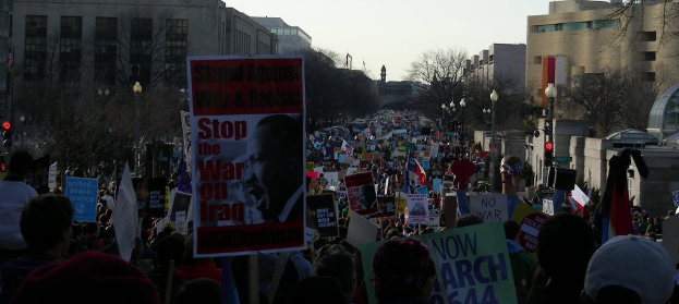 Eine Gruppe von Menschen, die auf einer Straße protestieren und Schilder halten, mit Gebäuden, Bäumen und Laternen im Hintergrund unter einem Himmel.