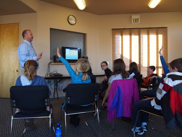 Ein Mann steht in einem Klassenzimmer und spricht zu einer Gruppe sitzender Kinder. Im Hintergrund sind ein Monitor, Lautsprecher, eine Uhr, ein Fenster und eine Tür zu sehen.