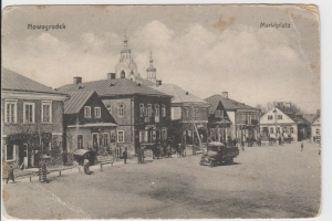 Ein Schwarz-Weiß-Foto einer Stadtstraße in Nowogrodeek, Deutschland, mit Gebäuden, Menschen, Karren, Pfählen, Bäumen und einem Himmel im Hintergrund, mit dem Text "Marktplatz in Nowogrodeek, Deutschland" oben und unten.