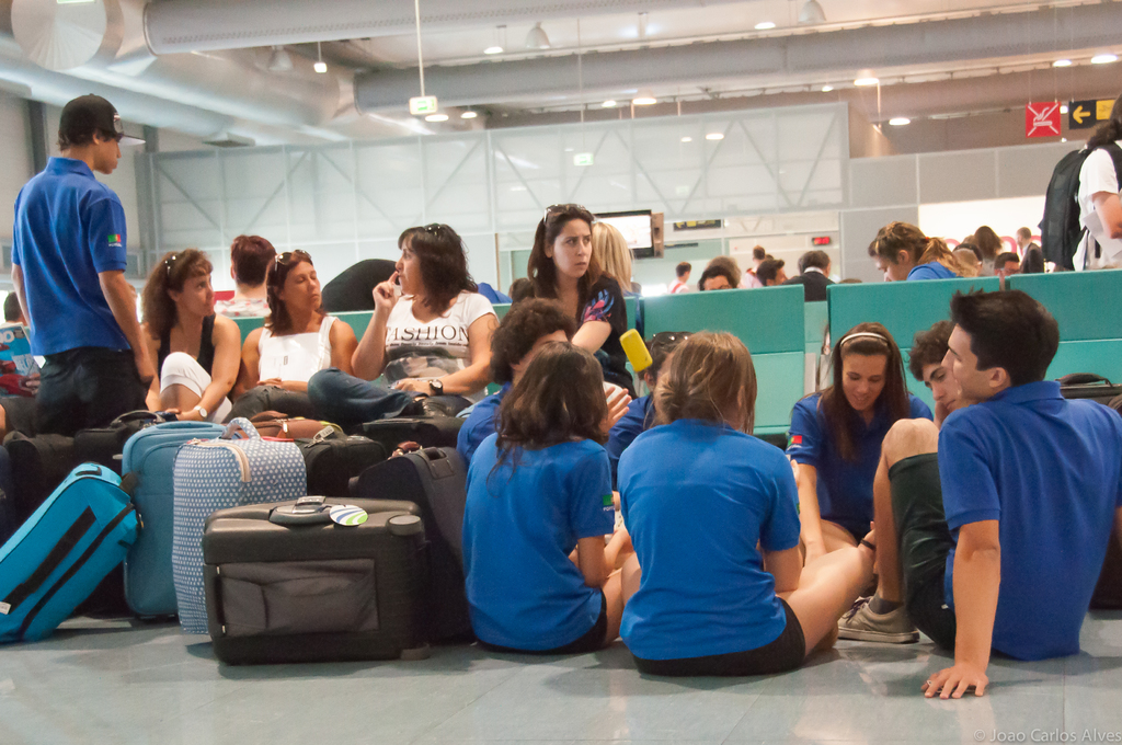 Ein belebter Flughafeninnenraum mit vielen Menschen, die auf ihre Flüge warten, Gepäckstücke links und Glastüren mit Hinweisschildern im Hintergrund.