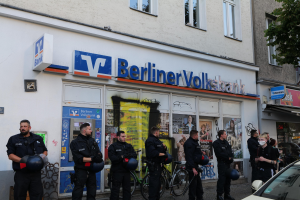 Eine Gruppe von Polizeibeamten vor einer Berliner Volksbank, einige mit Helmen, mit einem Auto auf der rechten Seite und einem Baum auf der linken Seite, vor einem Hintergrund von Gebäuden mit Fenstern und Texttafeln.