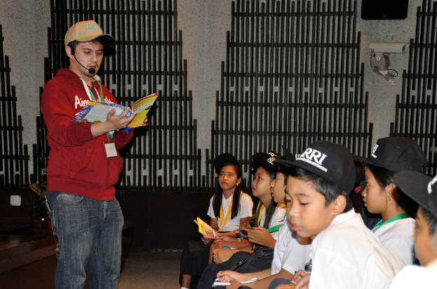 Ein Mann in einer marinefarbenen Kapuzenjacke und Jeans hält ein Buch und spricht in ein Mikrofon, während rechts ein paar Kinder in weißen T-Shirts und schwarzen Mützen zuhören; eine Kamera ist an der Wand im Hintergrund angebracht.
