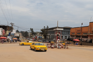 Eine belebte Stadtstra├če mit einem gelben Taxi, das fahrt, Passanten mit Schirmen auf den Gehwegen, Geb├Ąude mit Fenstern, Strommasten mit Kabeln und ein bew├Âlktes Himmel.