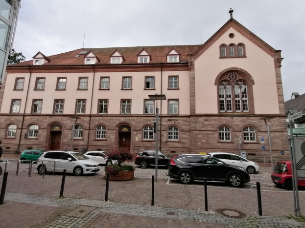 Ein großes Gebäude in Heidelberg, Deutschland, mit zahlreichen parkenden Autos davor, Straßenlaternen, fahrenden Fahrzeugen auf der Straße, Personen auf dem Gehweg, Topfpflanzen, Bäumen und einem bewölkten Himmel im Hintergrund, verfügbar zur Miete.