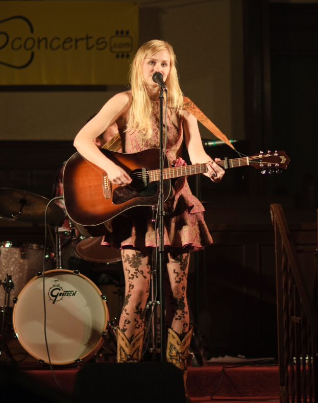 Eine Frau spielt Gitarre und singt auf der Bühne bei einem Konzert, mit einer Band und einem Bildschirm im Hintergrund und einem Podium unten.