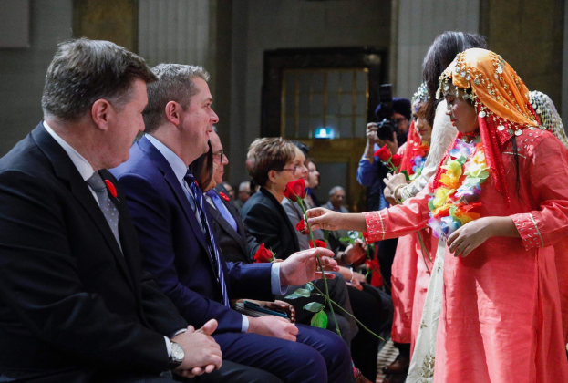Eine Gruppe von Menschen in einem Raum, mit einer Frau in einem Kranz und einem Bouquet in der Mitte, flankiert von einem Mann im Anzug und einer Person mit einer Kamera, vor einer Wand mit einer Tür.
