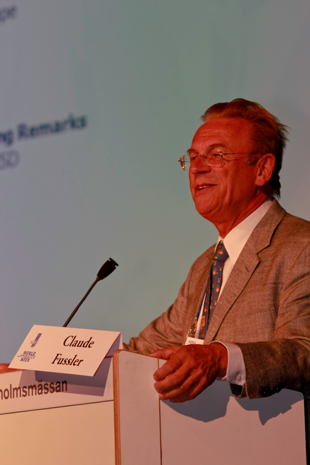 Ein Mann in einem weißen Hemd und einem grauen Blazer steht lachend vor einem Podium mit Mikrofon und Namensschildern, mit einem Projektionsschirm oder einer Banner im Hintergrund.