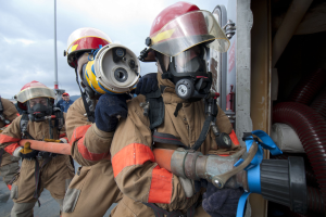 Feuerwehrleute in Schutzausrüstung, einer hält einen Schlauch, mit einer Stange und bewölktem Himmel im Hintergrund und Rohren auf der rechten Seite.