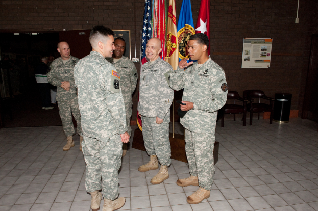 Funf Männer stehen und sprechen in einem Raum mit Stühlen, Flaggen, Tafeln, einer Wand und einer Tür im Hintergrund.