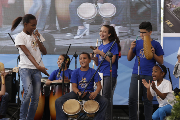 Eine Gruppe von Kindern, die auf einer Bühne Musikinstrumente spielen, mit einer Frau, die in der Mitte ein Mikrofon hält, umgeben von Mikrofonstativen, einem Banner im Hintergrund und Pflanzen auf der rechten Seite.