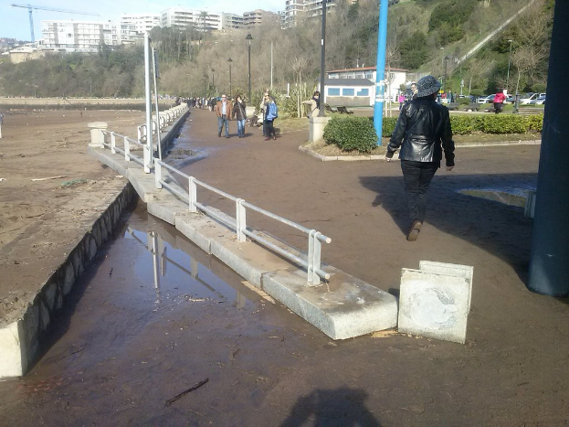 Eine Frau in einer schwarzen Lederjacke geht auf dem Boden neben einer Gruppe von Menschen in einem Fußgängerbereich, mit einem silbernen Geländer auf der rechten Seite und Bäumen und Gebäuden im Hintergrund.