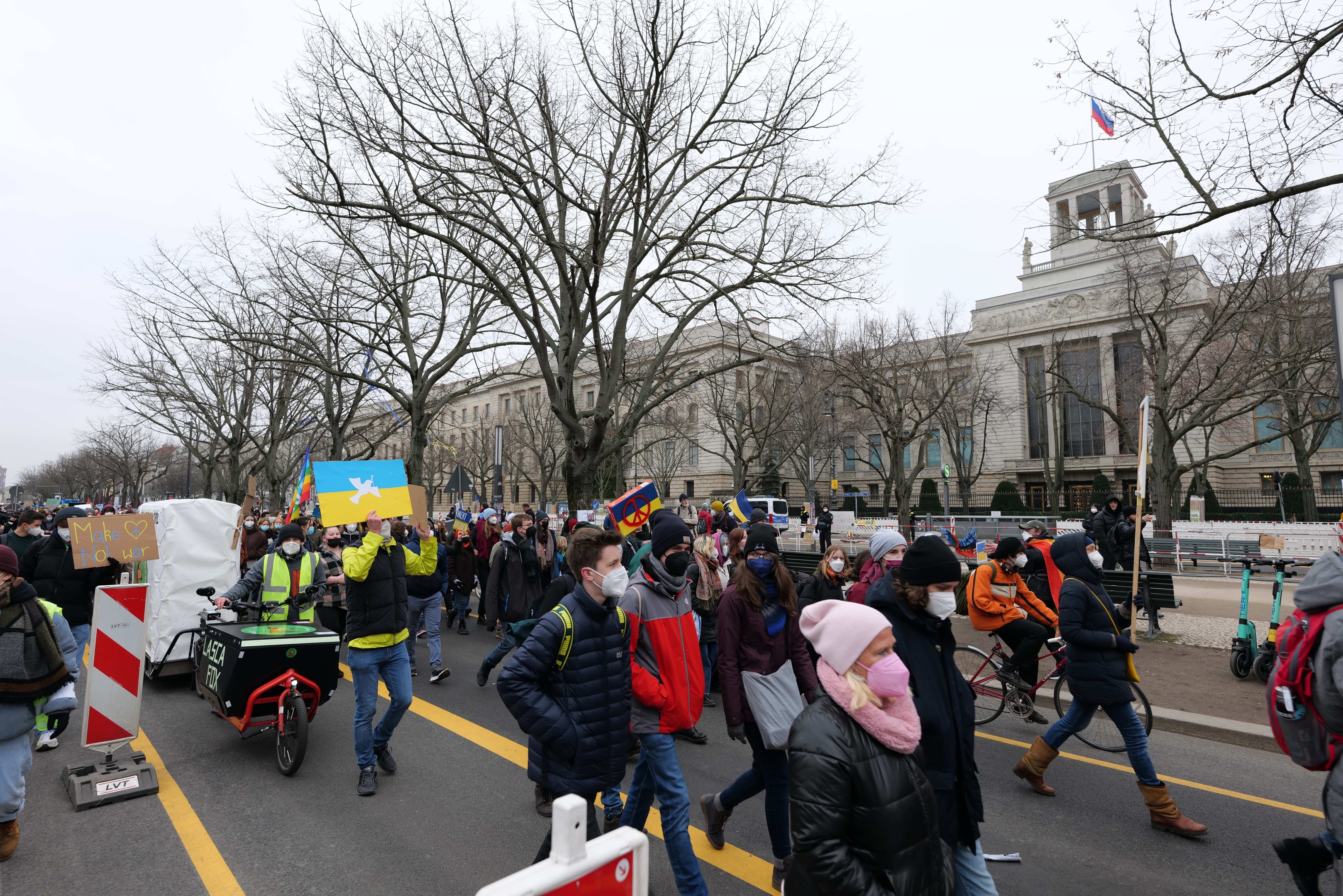Eine große Protestmarsch mit Menschen, die eine Stra√e in Washington, D.C. entlanggehen, einige halten Schilder und andere fahren Fahrröder, mit Bäumen, Schildern und einem klaren blauen Himmel im Hintergrund.