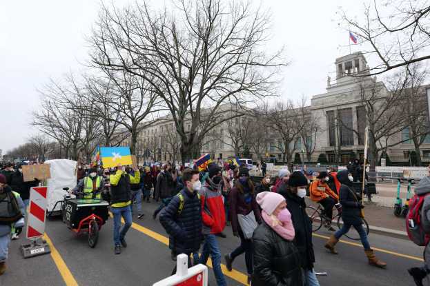 Eine große Protestmarsch mit Menschen, die eine Stra√e in Washington, D.C. entlanggehen, einige halten Schilder und andere fahren Fahrröder, mit Bäumen, Schildern und einem klaren blauen Himmel im Hintergrund.