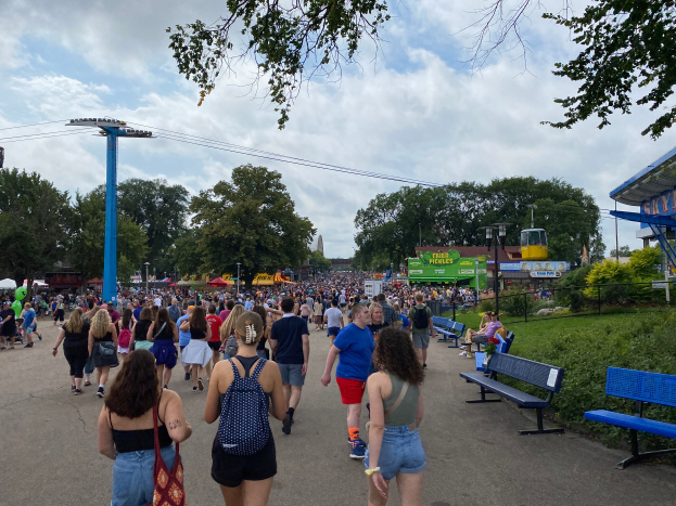 Eine Menschenmenge, die eine Straße mit Bänken, Pflanzen, Gras, Bäumen, Pfählen, Laternen, Kabeln und einem Schuppen entlanggeht, wobei einige Taschen tragen, unter einem bewölkten Himmel.