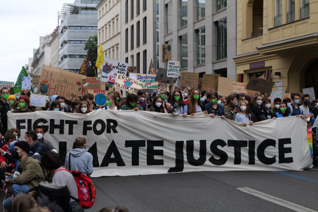 Eine große Gruppe von Menschen marschiert auf einer Straße, hält ein Banner mit der Aufschrift "Kampf für Hassjustiz", einige tragen Masken und halten Schilder, Gebäude, Bäume und einen klaren blauen Himmel im Hintergrund.
