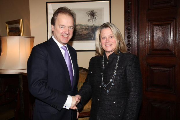 Eine Frau und ein Mann im Anzug lächeln, schütteln sich die Hände und sehen sich in einem Raum mit einer Wand, Fotorahmen, einer Lampe, einer Tür und ein paar Gegenständen an.