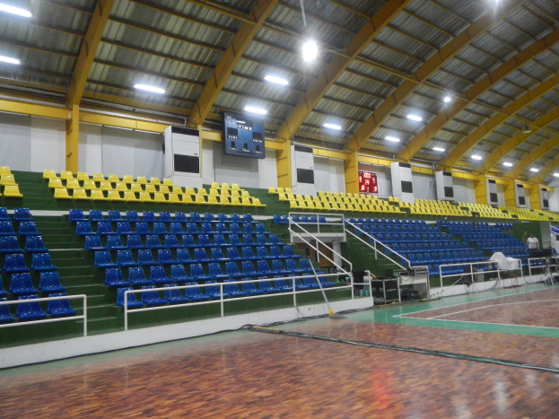 Großer Hallen-Sportplatz mit Holzboden, blauen und gelben Sitzplätzen, Geländern, Treppen zum Sitzbereich, wandmontierten Bildschirmen, Deckenleuchten und einigen Menschen.