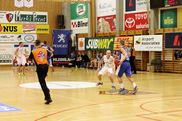 Ein Basketballfeld mit Spielern im Vordergrund und einer Holzwand mit Postern und Bannern im Hintergrund.