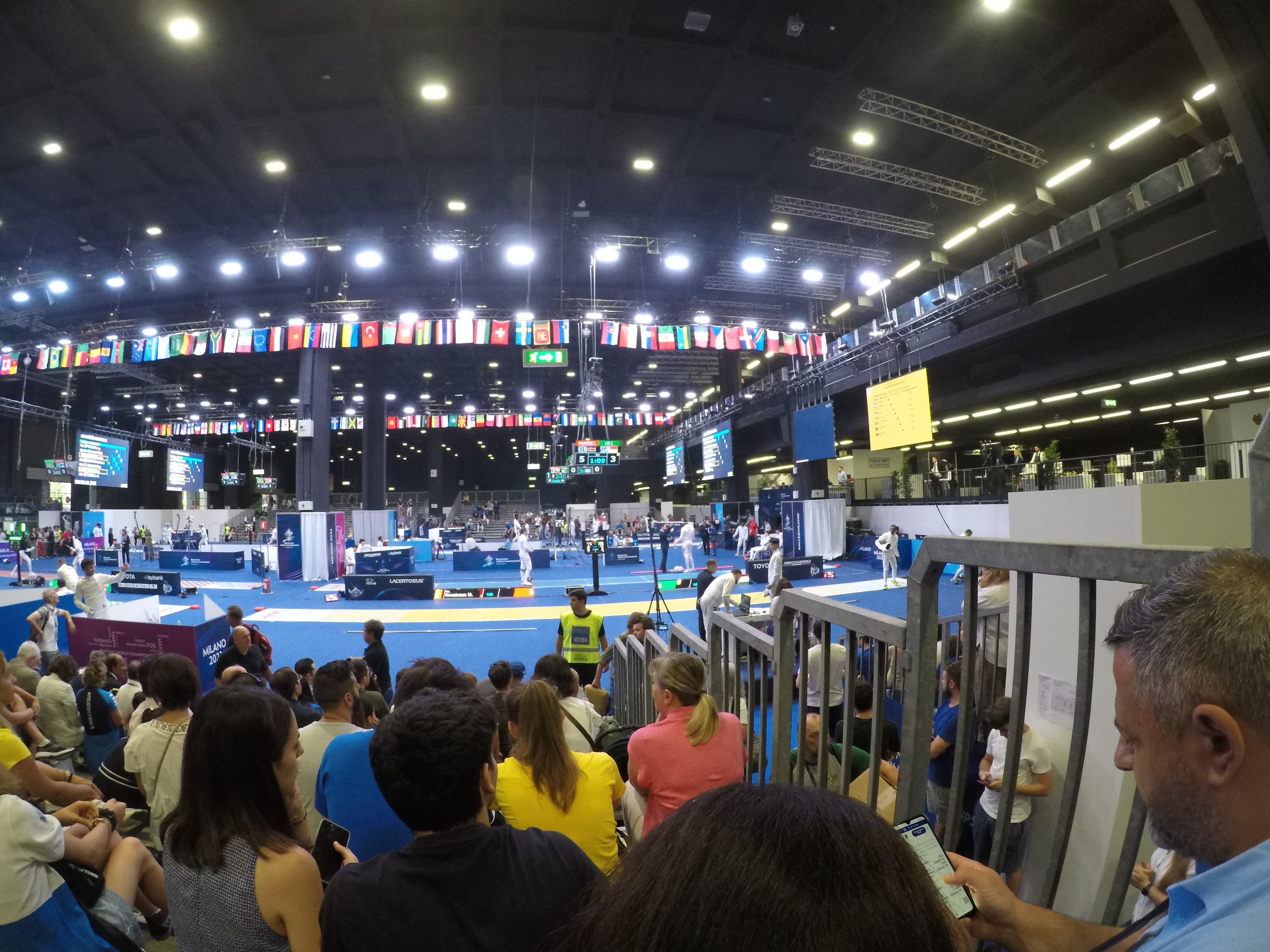 Eine große Menschenmenge sitzt und steht in einer Indoor-Sportarena mit Bildschirmen, Flaggen und Deckenbeleuchtung im Hintergrund.