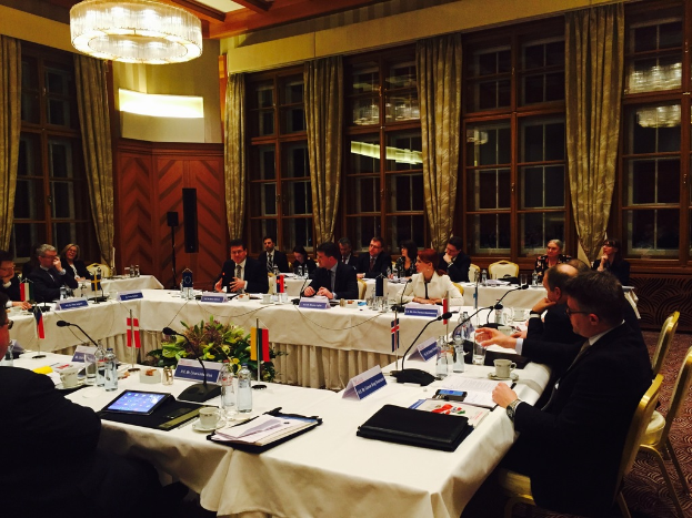 Menschen an einem Tisch mit verschiedenen Büromaterialien, darunter Ordner, Papiere, Getränke und elektronische Geräte, in einem Konferenzraum mit roter Wand, grünen Vorhängen und sichtbarem Decke.