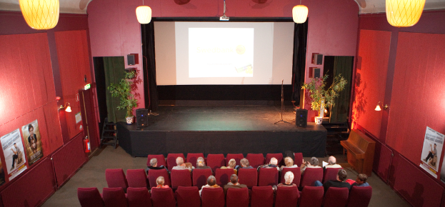 Ein Auditoriumsinneres mit einer Bühne, Leinwand, Lautsprechern, Pflanzen, Vorhang in der Mitte, Lampen und einem Projektor oben, einer Wand mit einem Plakat, Feuerlöscher, Lampen und Schildern links und Menschen, die auf Stühlen sitzen, unten.