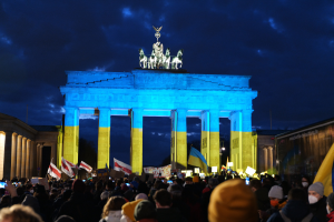 Eine Menschenmenge steht vor dem Brandenburger Tor in Berlin, Deutschland, mit Fahnen und Plakaten in den Händen, einem Banner auf der rechten Seite und dem Torbogen mit Statuen und Säulen unter einem bewölkten Himmel.