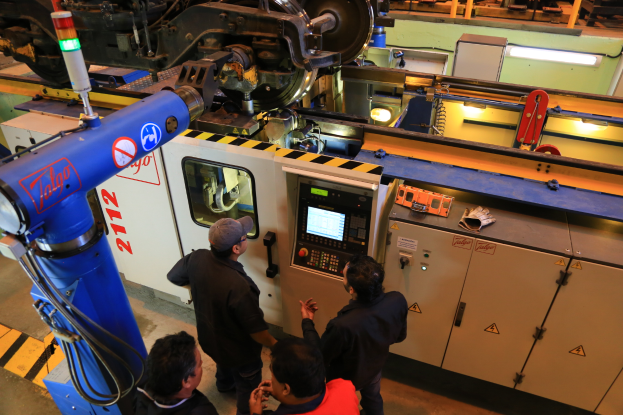 Vier Männer stehen um eine Maschine mit einem Bildschirm und Tasten in einer Fabrik, mit anderen Maschinen im Hintergrund.